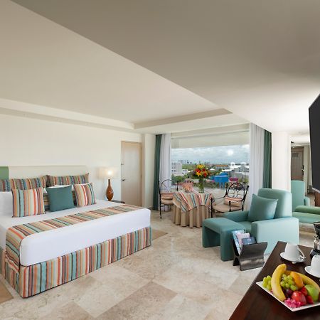 فندق غراند أويسيز بالم شامل كليًا كانكون المظهر الخارجي الصورة The photo shows a spacious hotel room with modern decor. It features a large bed with a striped duvet and decorative pillows, flanked by two lamps. There are comfortable seating areas with light green chairs and a small table. A balcony can be seen i