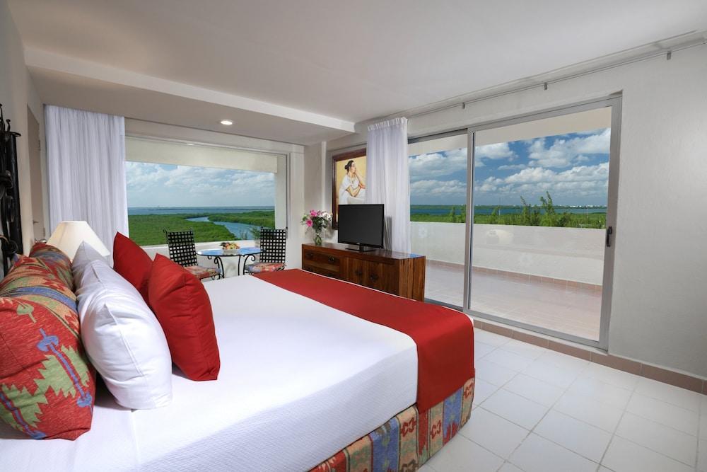 فندق غراند أويسيز بالم شامل كليًا كانكون المظهر الخارجي الصورة The photo shows a modern hotel room featuring a large bed with white linens and red accents. There are decorative pillows on the bed, and a bedside table with a lamp. To the side, there’s a large window with white curtains that offers a view of a bal