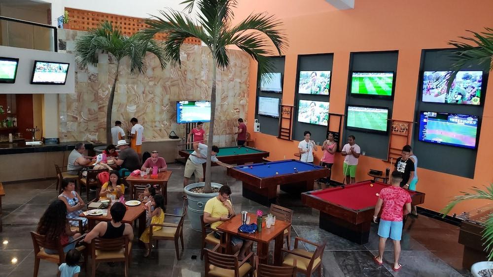 فندق غراند أويسيز بالم شامل كليًا كانكون المظهر الخارجي الصورة The photo shows a lively recreational area inside a building, likely a bar or game room. There are several pool tables, one blue and one red, where people are playing or waiting for their turn. 

Around the area, groups of people are seated at tables