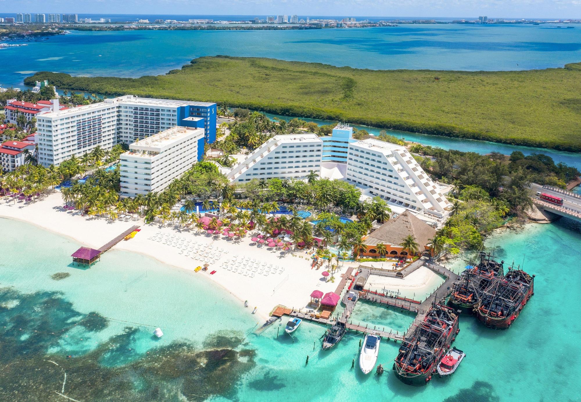 فندق غراند أويسيز بالم شامل كليًا كانكون المظهر الخارجي الصورة The photo shows a beautiful coastal resort area. In the foreground, there is a sandy beach with lounge chairs and umbrellas, while the clear turquoise waters are visible. The resort features large white and blue buildings, with lush greenery surround