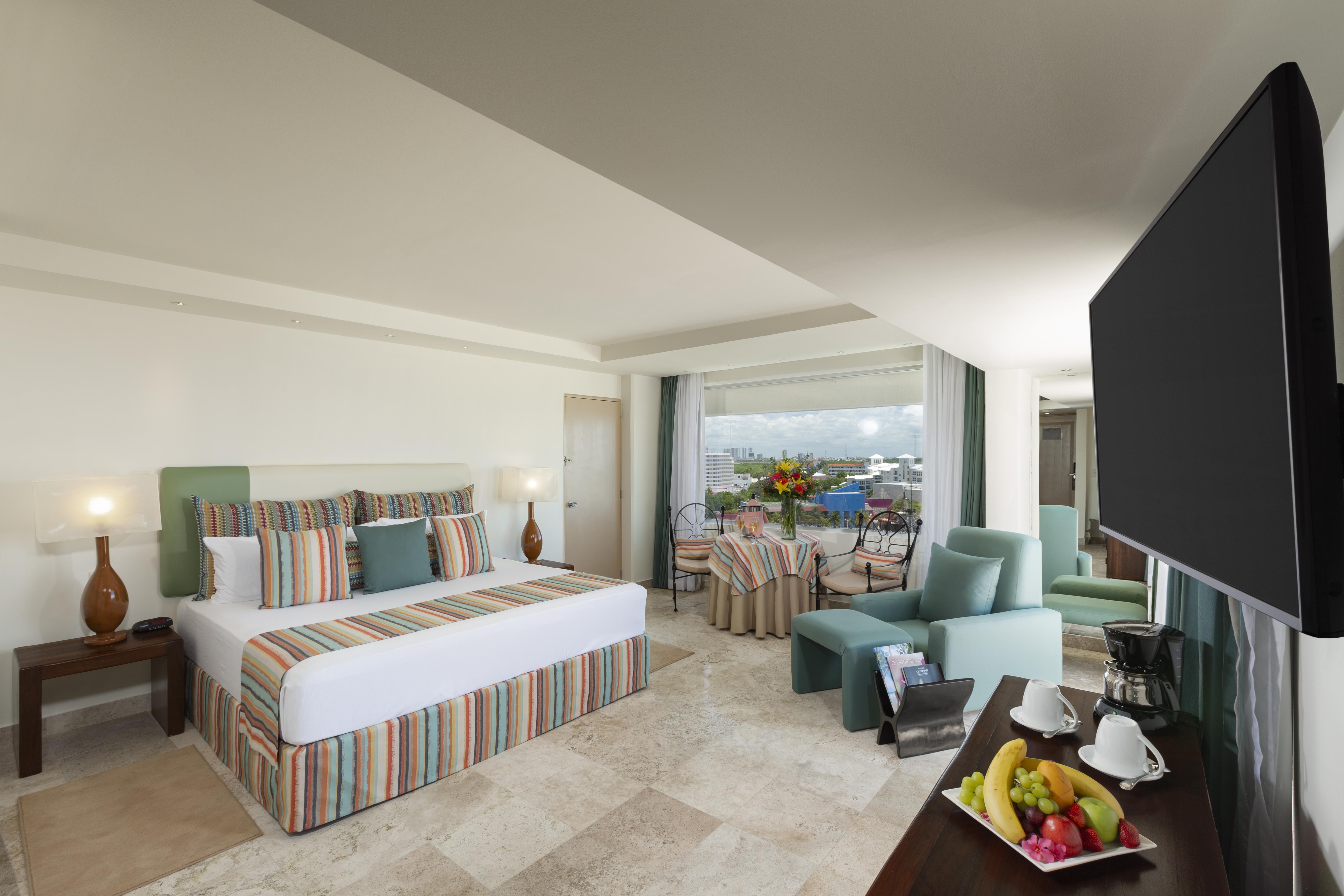 فندق غراند أويسيز بالم شامل كليًا كانكون المظهر الخارجي الصورة The photo shows a spacious hotel room with modern decor. It features a large bed with a striped duvet and decorative pillows, flanked by two lamps. There are comfortable seating areas with light green chairs and a small table. A balcony can be seen i