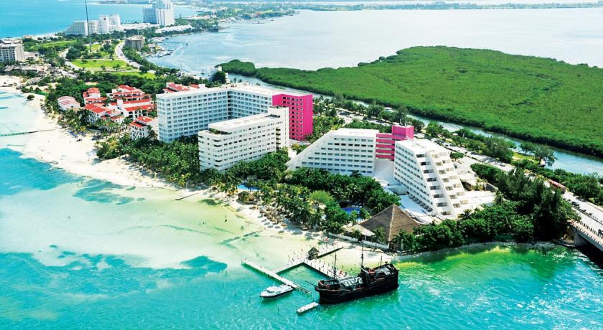 فندق غراند أويسيز بالم شامل كليًا كانكون المظهر الخارجي الصورة The photo shows a coastal resort area featuring several large hotel buildings, some of which are white and others are pink. The hotels are situated near a sandy beach, with clear blue water stretching out into the background. There are palm trees and