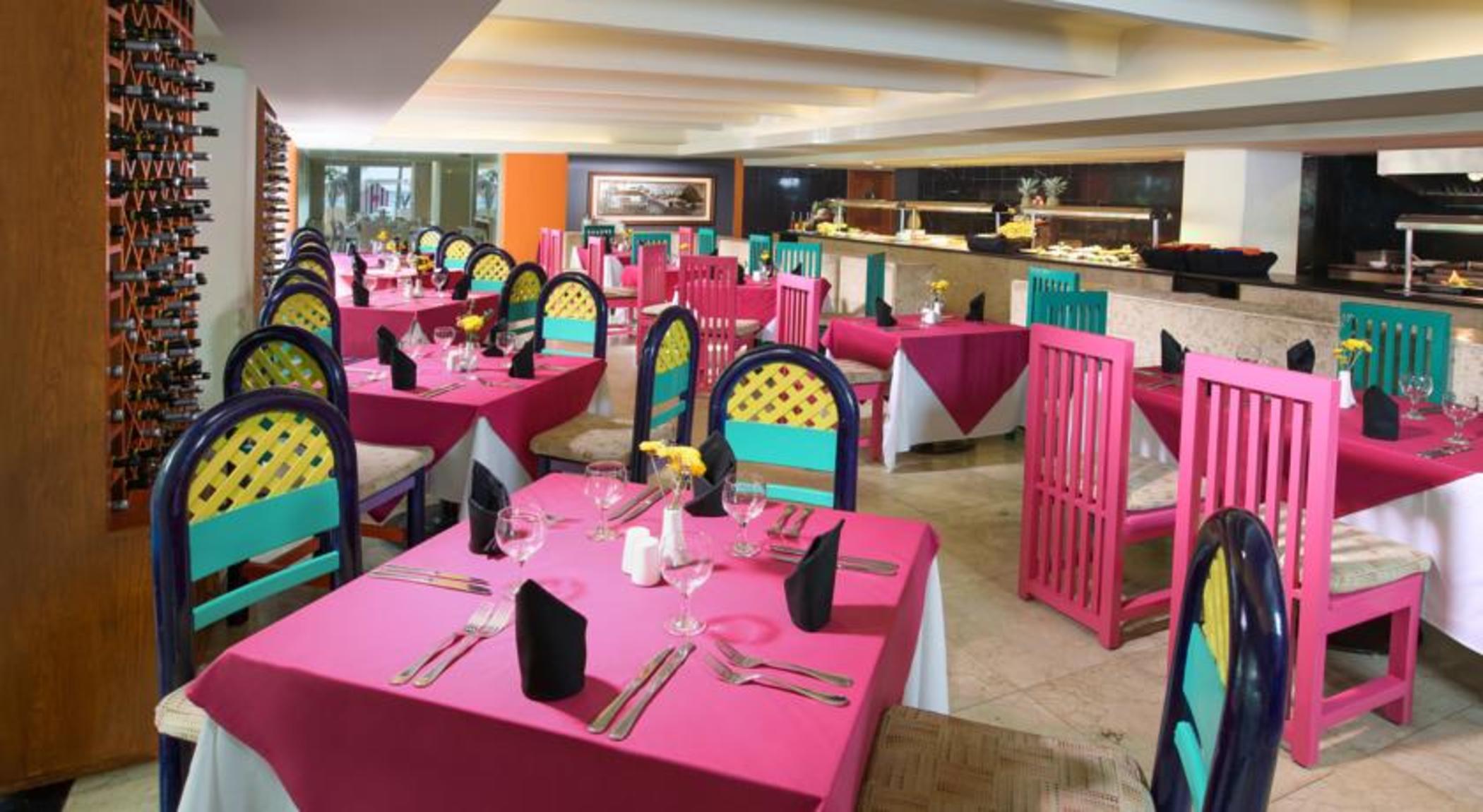 فندق غراند أويسيز بالم شامل كليًا كانكون المظهر الخارجي الصورة The photo shows a vibrant restaurant interior featuring colorful decor. There are multiple tables covered with bright pink tablecloths. Each table is equipped with utensils, napkins, and a small centerpiece, possibly a flower. The chairs are painted 