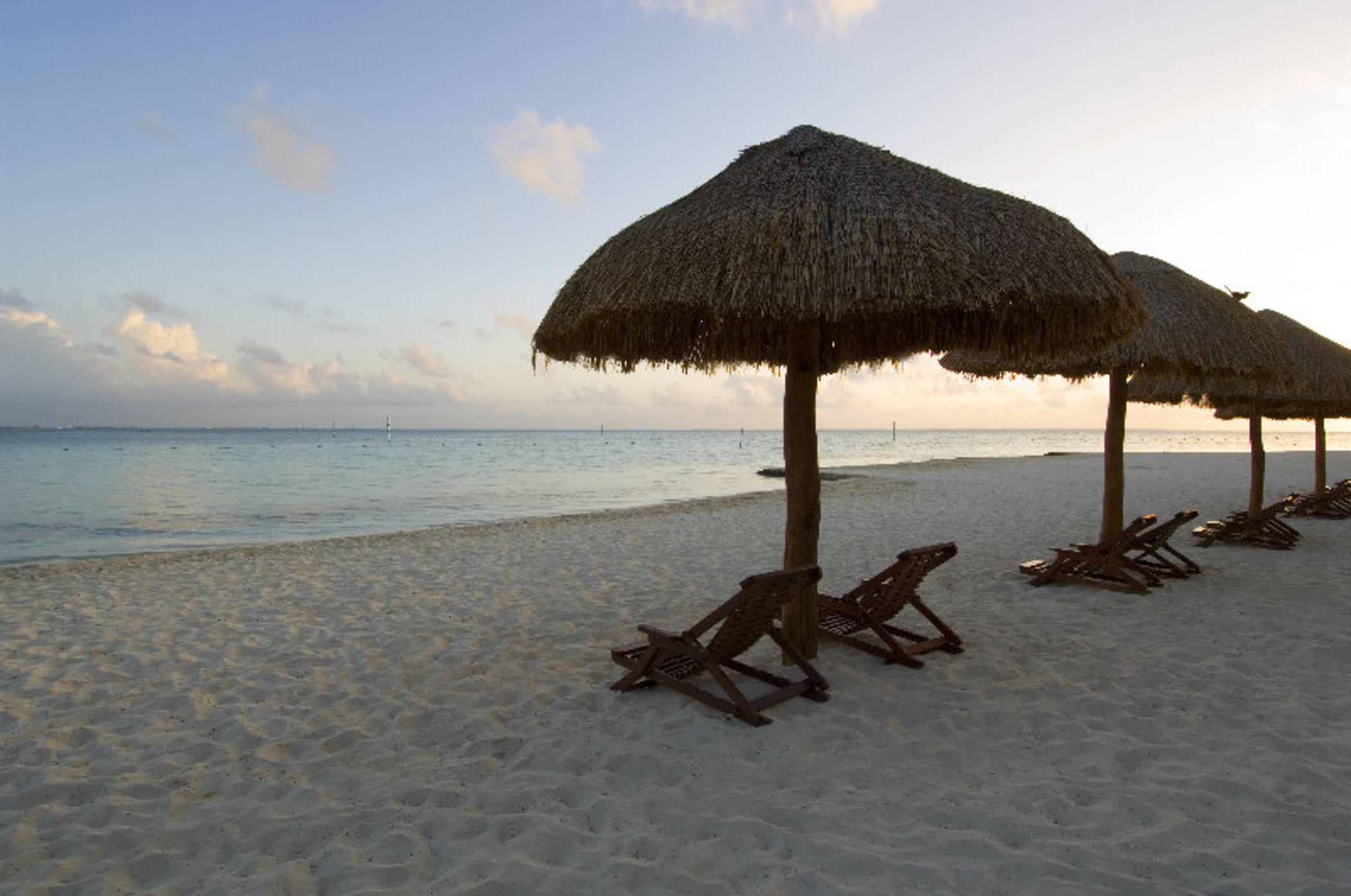 فندق غراند أويسيز بالم شامل كليًا كانكون المظهر الخارجي الصورة The photo depicts a serene beach scene featuring several thatched umbrellas on the sand. Beneath the umbrellas are wooden lounge chairs, positioned for relaxation. The beach has soft, white sand, and the calm waters of the sea reflect the colors of t