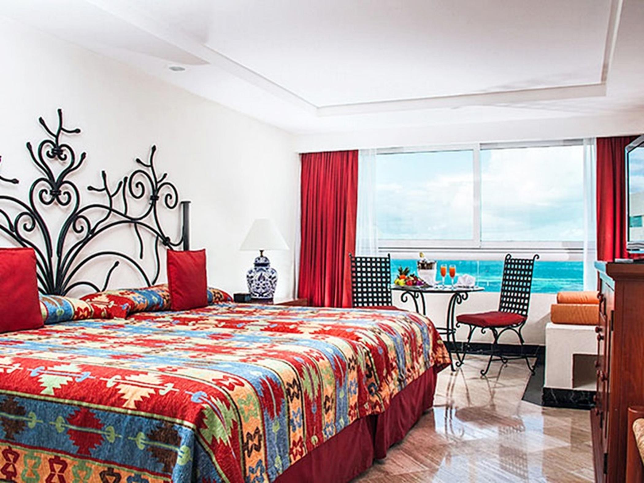 فندق غراند أويسيز بالم شامل كليًا كانكون المظهر الخارجي الصورة The photo shows a well-decorated hotel room featuring two large beds with vibrant, patterned bedding. The headboards are stylishly designed, and the room has colorful accents like red curtains and cushions. A small table with chairs is positioned by 